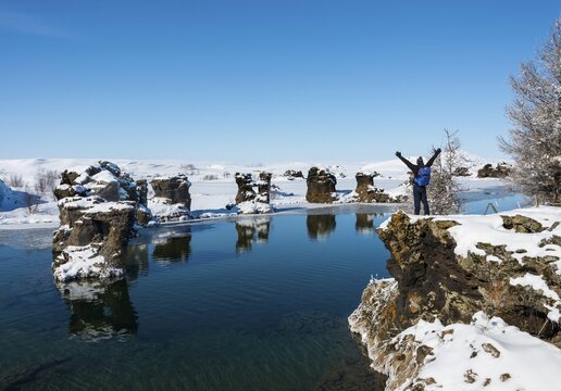 Young man stretches his arms into the air, view of volcanic rock formation in the water, Lake M&yacute;vatn in winter, H&ouml;f&eth;i Peninsula, Iceland