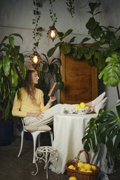 A woman sits at a round table surrounded by plants, reading a book. She has her feet relaxed on the table. A basket of lemons sits nearby. The space has warm light and a calm atmosphere
