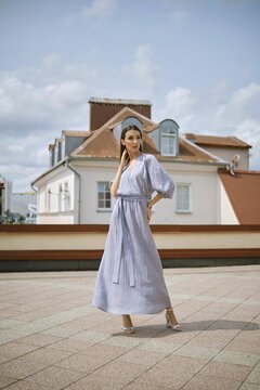Graceful woman stands on a rooftop, wearing a long, light blue dress with puffed sleeves. She poses elegantly against a backdrop of cloudy skies and rooftops, showcasing her fashion sense