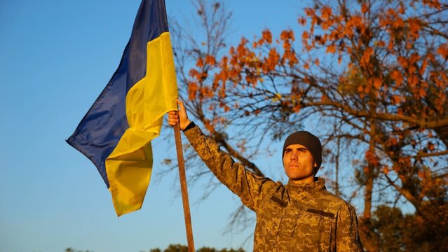 Male ukrainian army soldier lifting blue-yellow banner in honor of victory against russian aggression. Young military in camouflage uniform raised flag of Ukraine at countryside. Invasion resistance