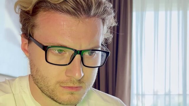 Close-Up of a Young Man Working on a Laptop, Wearing Glasses with the Screen Light Reflecting in His Lenses