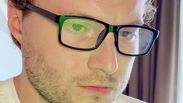 Close-Up of a Young Man Working on a Laptop, Wearing Glasses with the Screen Light Reflecting in His Lenses