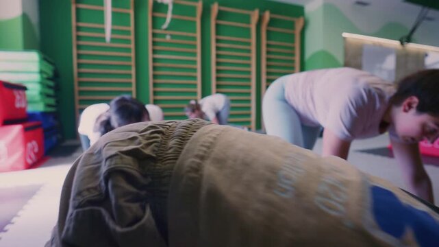 Children and an adult participate in a fitness class, performing crawling exercises on the gym floor. Young kids engage in dynamic physical activity led by a trainer in a modern sports facility.