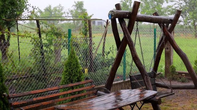 A cozy wooden swing, table, and bench get wet under a pouring summer rain in a rural garden or backyard. The furniture surface glistens with moisture, surrounded by green trees, tuja bushes, and a mes