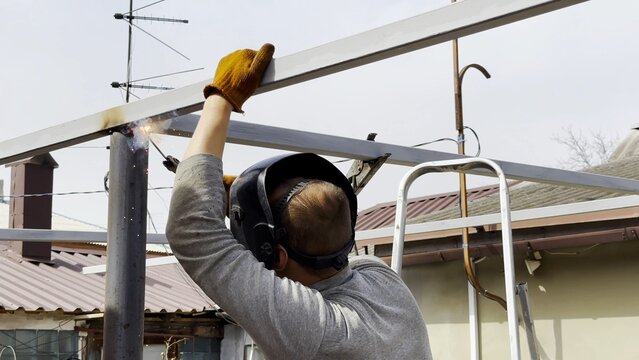 Manual laborer in protective mask weld iron detail on carport frame. Professional young repairman with welding machine working at construction. Male welder operating with metal at the site. Close up