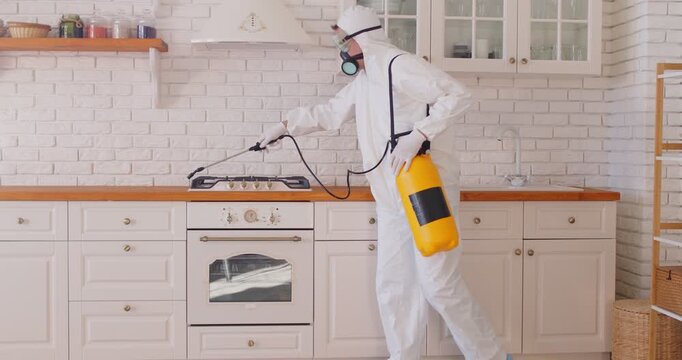 Exterminator holding sprayer with nozzle and canister of insecticide for kitchen disinfecting. Pest control worker in protective mask and suit spraying stove with pesticide for insect cleaning