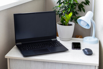 Fototapeta premium Modern home office workspace with a black laptop, smartphone, wireless mouse, blue desk lamp, and green potted plants on a light wooden table. Interior