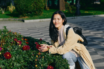 Fototapeta premium A woman kneeling beside vibrant flowers in a garden, smiling softly as she admires blossoms, wearing a light coat and backpack, enjoying a sunny outdoor moment.