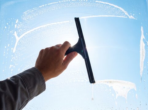 Hand using black squeegee to clean soapy window against blue sky