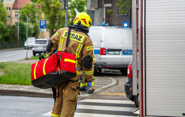 A firefighter in a beige protective suit with the word STRAŻ and a yellow helmet seen from behind, carrying a large red medical kit bag on his shoulder across a pedestrian zebra crossing. © Adam