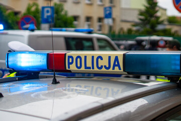 Blue police lightbar with the inscription POLICJA mounted on the roof of a silver patrol car flashing bright LED emergency lights against a blurred building and tree. © Adam