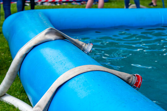 Close up of a blue portable water tank with fire hoses placed over the edge, metal and red coupling connectors, green grass and blurred legs in the background.
