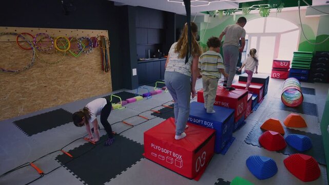 Young children are actively participating in a fun fitness session with various exercises. They are seen jumping and stepping on colorful soft boxes and hurdles in a modern gym.