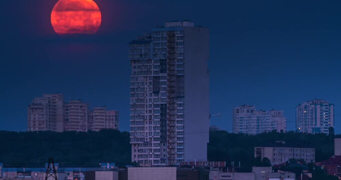 Large red full moon setting down behind modern skyscraper in Kyiv at night. Beautiful Strawberry Moon celestial event over the city skyline, Kyiv, Ukraine.