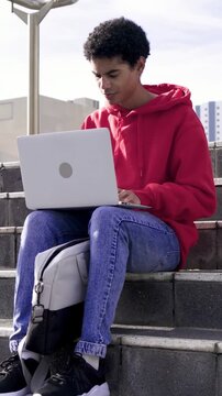 Vertical video: Settling on steps, man in red hoodie opening laptop, typing to study with backpack