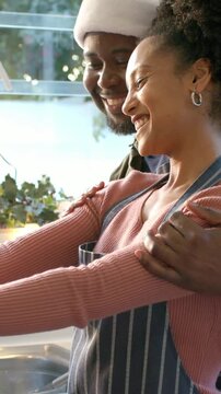 Vertical video: Reaching behind her, man hugging as woman rinsing glass in apron preparing meal