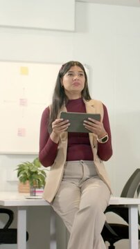 Vertical video: Glancing at coworker gesturing right, woman in maroon top holding tablet at table
