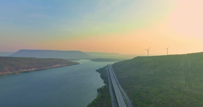 Aerial view of Lam Takhong Dam at twilight showing bright highway lights curving along the calm blue lake, surrounded by mountains, wind turbines, and scenic nature under a peaceful evening sky.
