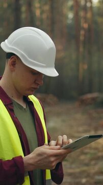 A forestry engineer, also a lumberjack, wearing a safety helmet and bright vest, examines a touchscreen tablet at a forest work site, surrounded by trees, planning tasks on the device's screen