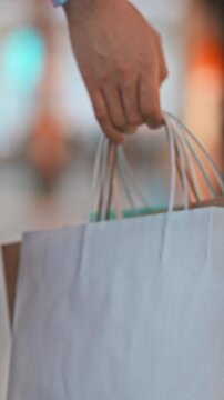 A girl's hand grips paper-packaged shopping bags filled with items as she walks through a bustling shopping centre, the scene showing a typical consumer purchase activity in a retail environment