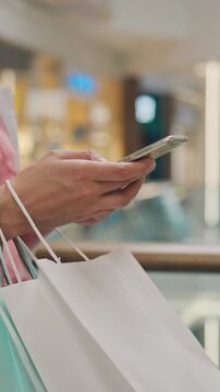 A girl's hands clutch a smartphone and several paper shopping bags while standing in a busy shopping centre, she stops briefly, eyes on the phone, the bags full of recent purchases resting beside her
