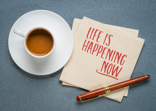 Life Is Happening Now. Inspirational handwritten note on a napkin with coffee and pen, encouraging mindfulness, living in the present, and embracing every moment.