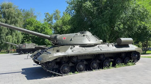 Military Machine: A powerful green tank, a symbol of historical conflict and defense, stands sentinel on a paved surface, surrounded by lush greenery and dappled sunlight.