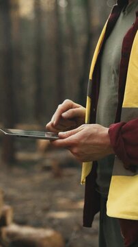 A senior logger engineer in a bright safety vest and helmet stands amid dense green trees, eyes fixed on a tablet screen as he monitors and directs the timber-cutting operation, field digital control