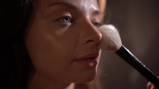 Professional makeup artist applying loose powder with large fluffy brush on model's face in close-up, focus shift between brush and face, warm back light, creating flawless matte finish.

