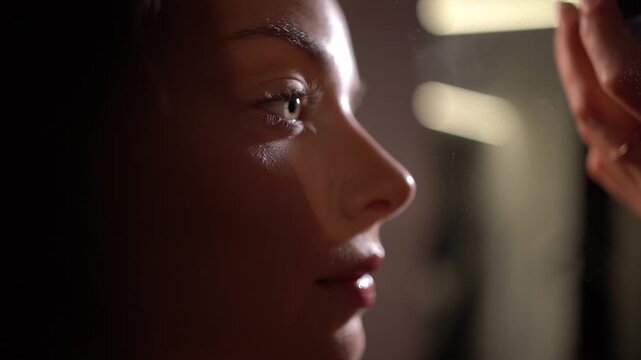Professional makeup artist applying loose powder with fluffy brush on model's face in extreme close-up, profile view, model closing eyes with slight smile, floating powder particles in back light.