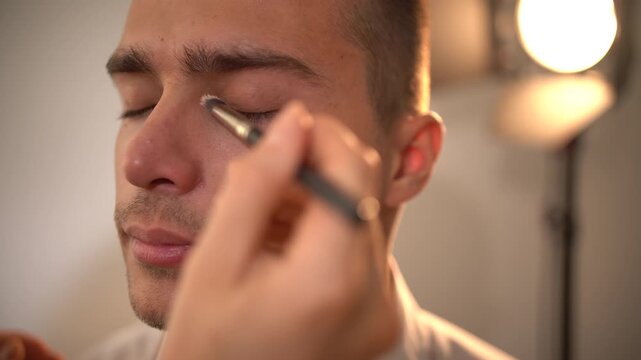 Professional makeup artist woman using small brush to apply concealer to handsome young man's right side of face in profile view, model's eyes closed throughout, artist's hand and brush visible.
