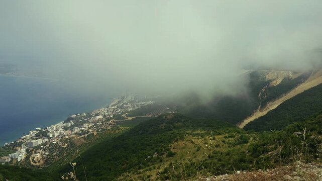 Thick Clouds Covering Mountain Slopes Near Sea Coast.  Foggy Mist Over Green Hills And Coastal Town Buildings