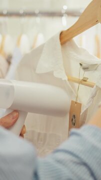 An Asian store worker girl carefully uses a modern steamer on hanging clothes, smoothing fabric wrinkles in a retail area, close-up shows her hands, the steamer nozzle and the textured textiles