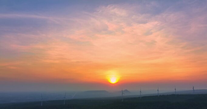 Aerial view of Lam Takhong Dam at sunrise. where soft golden light spreads across the lake and hills. The elegant wind turbines stand tall, turning gently in the morning breeze amid a sky 
