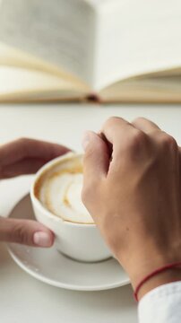 At a white restaurant table, a young girl holds a cappuccino cup, stirs the hot coffee with her hands, then drinks it. The bright dining area has white decor and soft ambient light