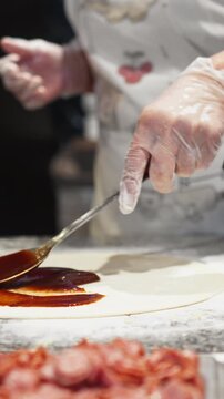 Chef prepares street-food pizza, spreading bright red chili sauce over rolled dough on metal table, hands steady as dough is coated before baking, highlighting the pizza cooking process at busy stall