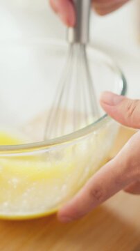 Close-up shows a woman's hands gripping a whisk, beating raw yellow eggs in a clear glass bowl under bright sunlight, capturing the mixing step of a breakfast preparation process in a kitchen