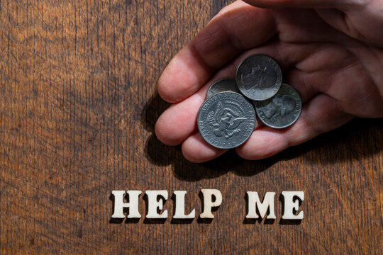 Hand holding coins over help me text on wooden background. Closeup of human hand holding US dollar coins above wooden letters spelling help me on dark desk representing financial crisis and poverty