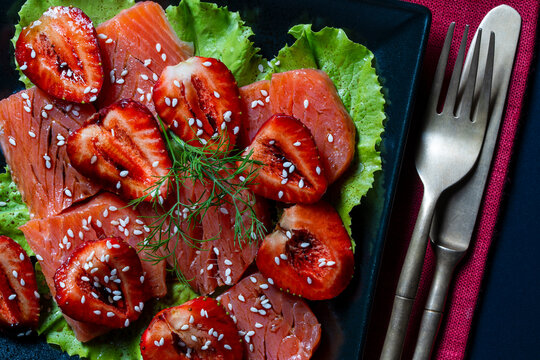Salmon carpaccio with fresh strawberries and sesame seeds. Gourmet salmon carpaccio paired with sweet strawberry halves on green lettuce leaves with sesame seeds served on black plate