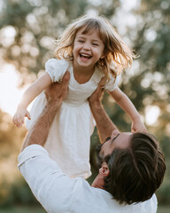 Fototapeta premium Sunlight bathes a joyful child, arms outstretched. Man lifts her high, laughing with pure delight. Golden hair swirls in the breeze, free and bright. Gentle touch, tender moment, care in motion