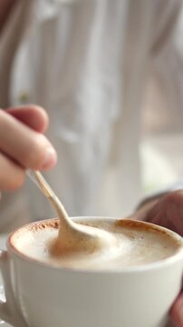 Girl in white clothes stirs barista-made cappuccino with a metal spoon, pushing the foam in a white cup while she eats lunch at a bright, sunlit cafe, the scene showing a typical coffee-break routine