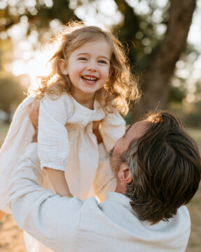 Golden light bathes a joyful child's face. Curly hair gleams as she laughs. Man lifts her with a tender smile. Sunset glow frames their happy moment. Perfect for family, parenting, or joyful moments