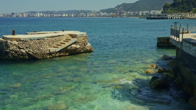 Old Stone Pier With Stairs In Clear Turquoise Sea Water Against Coastal City Buildings And Mountains On Sunny Day