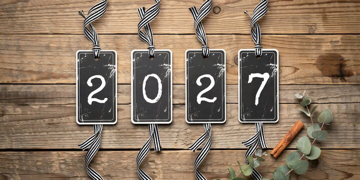 Four rustic black rectangular tags featuring white hand-painted numbers forming 2027 suspended by striped black and white ribbons against a weathered vintage brown wooden background