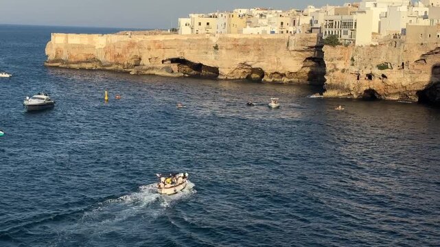 Small boats cruising along the limestone coast of Polignano a Mare, Puglia, Italy on the calm Adriatic Sea with historic waterfront in warm evening light. Summer travel, tourism, vacation, coastal lan