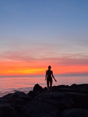 Fototapeta premium Silhouette of a woman standing on the rocks watching an incredible vibrant orange sunset over the sea.