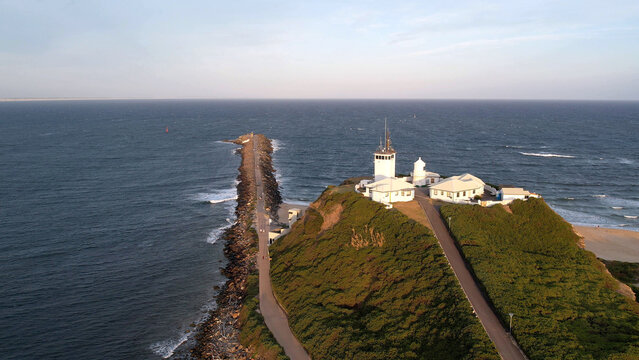 Aerial view of Nobbys Lighthouse situated on a grassy headland with a long breakwater extending into the ocean under a clear sky in Newcastle, New South Wales, Australia.