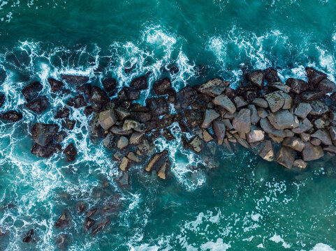 Aerial view of dark rocks forming a breakwater in the turquoise ocean with white sea foam and crashing waves in Marina del Rey, California, United States.