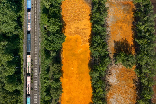 Aerial view of a freight train traveling on tracks alongside a vibrant orange mineral-rich sediment basin bordered by lush green forest.