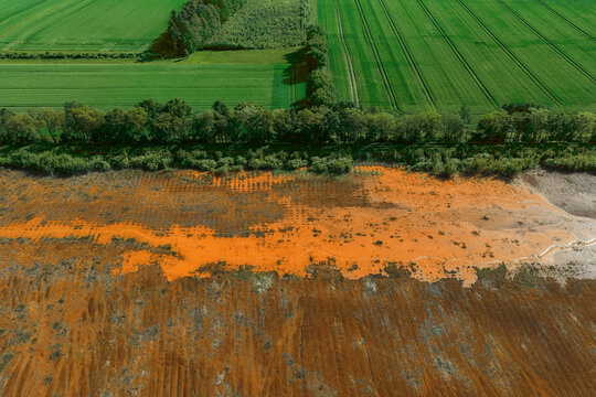 Aerial view of a striking contrast between lush green agricultural fields and a patch of orange-colored mineral-rich soil, separated by a dense line of trees.